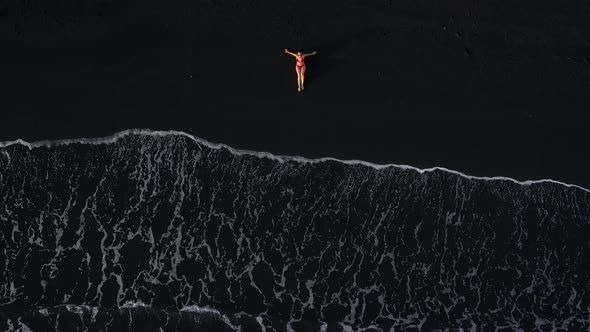 Top View of a Girl in a Red Swimsuit Lying on a Black Beach on the Surf Line alt