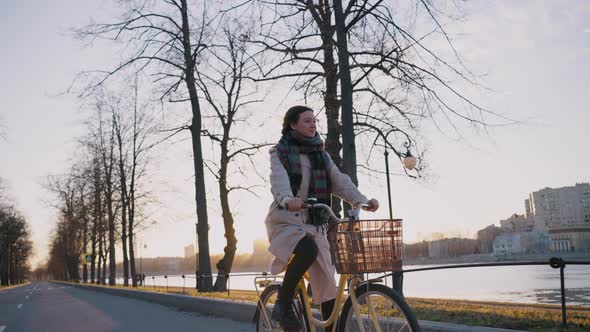 Cinematic Shot of a Beautiful Woman in a Raincoat Rides a Bike Along the Embankment Against the alt