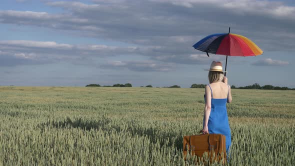 Girl in blue dress with suitcase and umbrella in wheat field alt