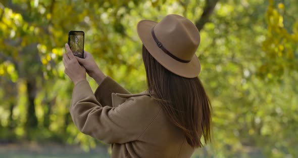 Pretty Brunette Caucasian Girl in Brown Hat and Coat Taking Photos of Autumn Park on Her Smart Phone alt
