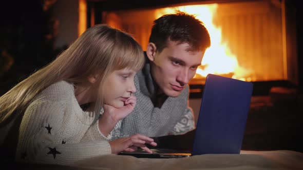 Children Play Together on a Laptop, Lie on the Floor Near the Fireplace alt