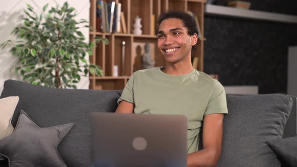 Cheerful Multiracial Man Wearing Casual Wear Sitting on the Sofa and Using Laptop alt