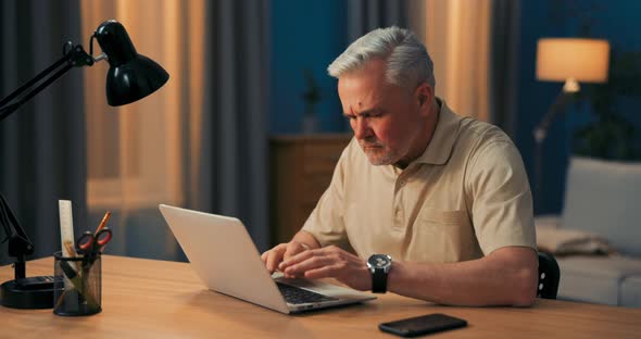 Elderly Man with Gray Hair Works on Laptop at Desk and Lamp in Evening alt