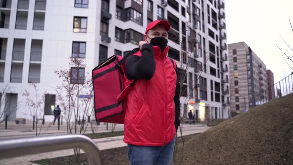 Portrait of Delivery Guy Wearing Red Uniform and Protective Mask with Thermal Bag Modern Buildings