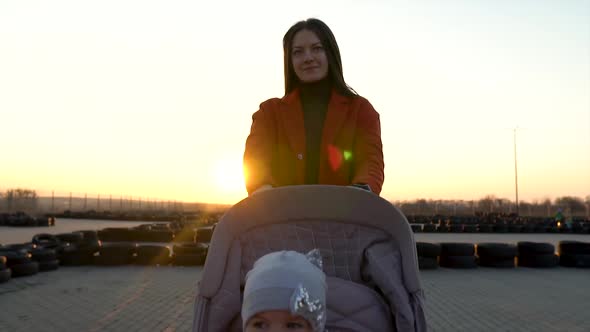 A Slow Motion of Confident Mother Walking with a Pram. Sunset Behind, Silhouette. A Little Toddler