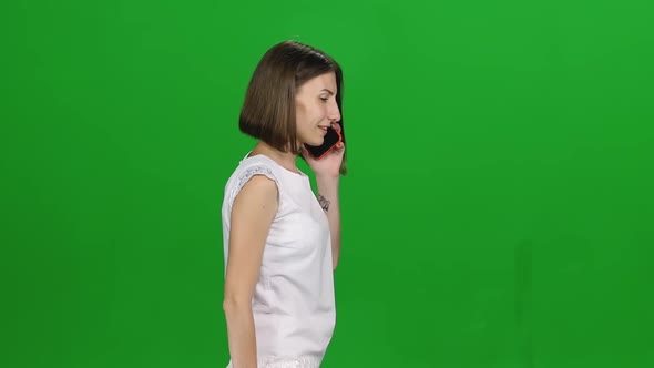 Portrait of Young Woman Is Calmly Walking, Talking on Mobile Phone at Green Screen. Profile View alt