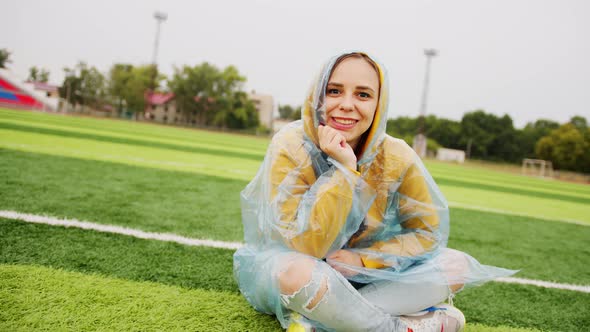 Young Woman in Raincoat Sitting on Football Field in Rainy Weather alt