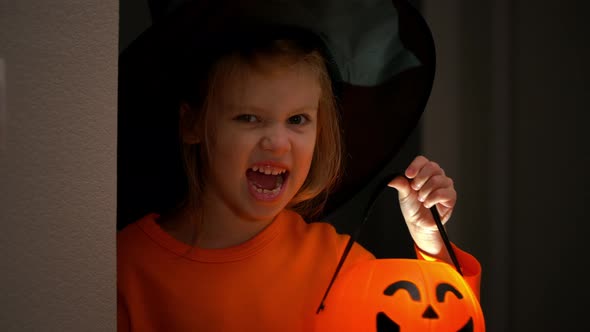 Little Girl in Witch Costume Fun Hides and Scares From Corner with Lantern Halloween Pumpkin in Dark