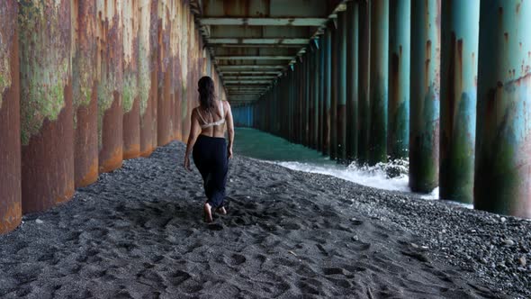 a Barefoot Woman Dances Under the Pillars of the Bridge Against the Background of the Incoming Waves alt