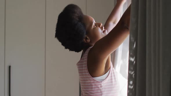 Happy african american woman widening curtains in bedroom alt