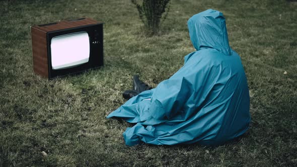 Woman in Blue Raincoat with Hood are Sitting on Grass and Watching Old Retro TV