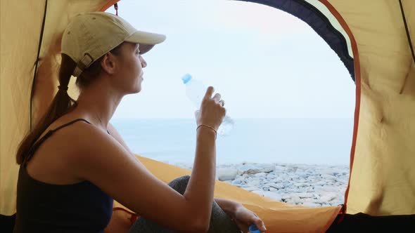 Woman is Sitting in Camping Tent Drinking Water From Bottle and Looking at Sea