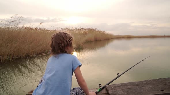 Little Girl Sits on a Bridge By the River Takes Out a Fishing Rod Without Fish alt