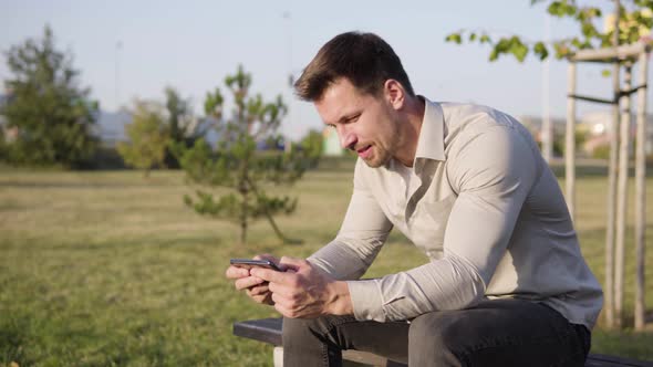 A Young Caucasian Man Plays a Game on a Smartphone As He Sits in a Park in an Urban Area alt