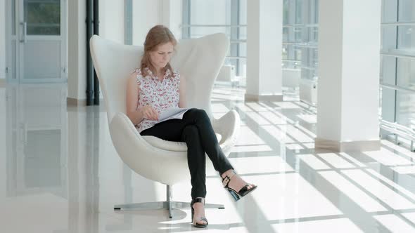 Attractive Businesswoman Sitting on a White Chair in a Glass Office and Checks Documents