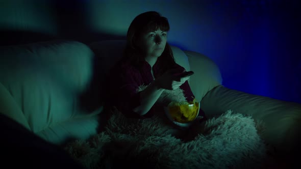 Brunette Woman Sits in Sofa with Chips at Night and Watches Tv with Emotion Switches Channels