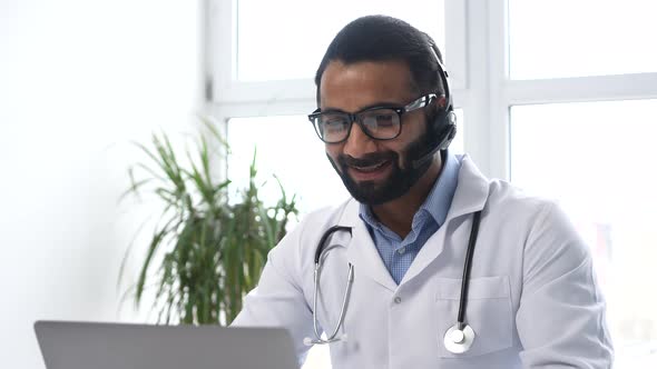 Indian Male Doctor with a Stethoscope in Gown Sitting at the Desk and Looking at the Laptop alt