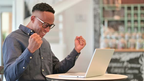 Excited African Man Celebrating Success on Laptop in Cafe  alt