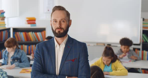 Young Male Teacher Standing in Classroom with Kids Sitting at Desk on Background alt