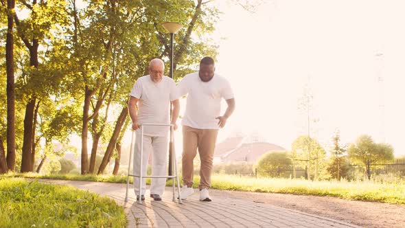 African-American caregiver is teaching disabled old man to walk with walker. Nurse and patient. alt