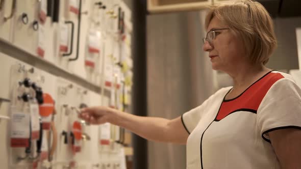 Elderly Woman is Examining Accessories for Furniture Fixed on a Stand in Shop alt