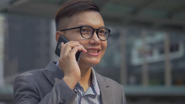 Asian men with glasses use phones in street near big office buildings.