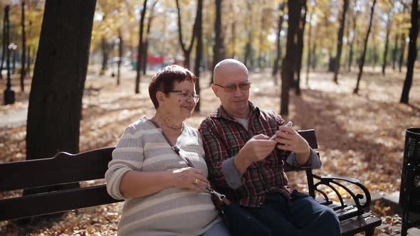 An Elderly Couple in the Autumn Park Sitting on the Bench Taking Selfies on a Smartphone