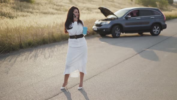Business Woman Holds a Cup of Tea and Talks on Phone at Broken Car Out of City alt