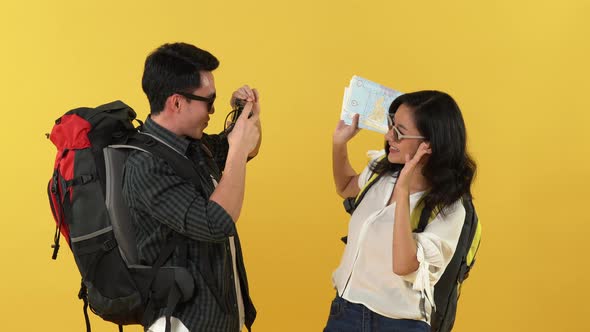 Asian tourist with backpack taking picture of his girlfriend with camera on yellow background