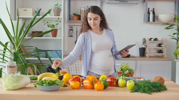A Pregnant Woman Looks for a Recipe on a Tablet And Makes a Salad in a Bright alt