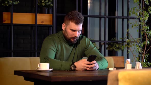 Young Bearded Hipster Man Sits at Table in Cafe,uses a Smartphone and Drinking Coffee