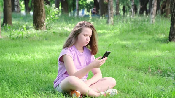 Teen Girl Holding Cell Phone on the Lawn in a City Park and Typing on Smartphone