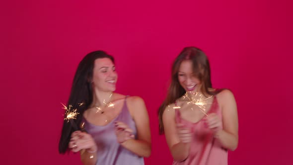 Two Happy Young Women Have Fun Dancing with Burning Sparklers on Red Background