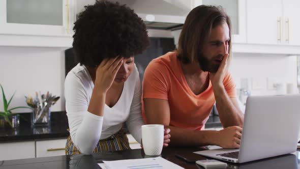 Stressed mixed race couple using laptop and calculating finances in the kitchen at home alt