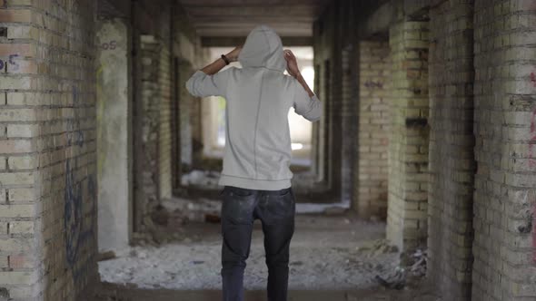Back View of Young Male Bandit Walking Along Abandoned Ruins and Putting on Hood. Wide Shot of alt