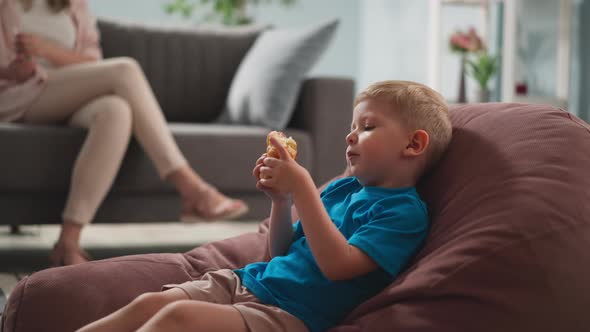 Little Child Sits in Bright Room on Armchair Eats Sandwich Next to His Mother Sitting on Couch alt