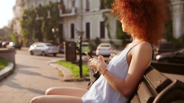 Attractive Readhead Girl Resting Looking at Phone Sitting on Bench Outside alt