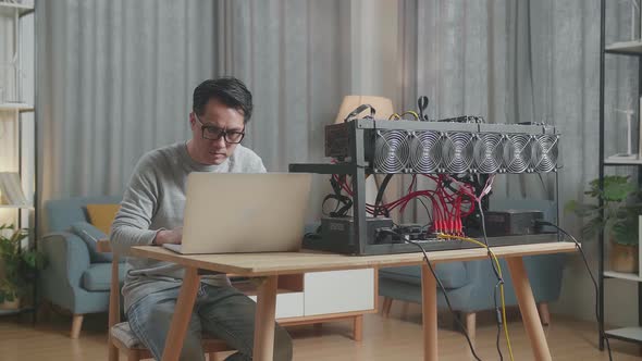 Serious Asian Man Wearing Glasses Using Laptop And Mining Rig For Mining Cryptocurrency alt