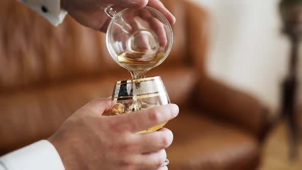 Closeup Male Caucasian Hands Pouring Drink From a Cup Into Cognac Glass alt