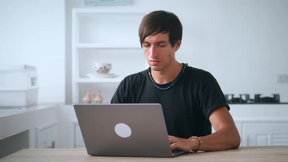 Young Man Freelancer Working at Laptop While Sitting at Kitchen Table at Home alt