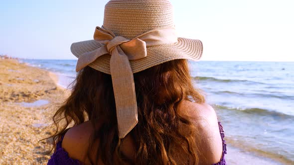 An Unrecognizable Girl with Brown Hair in a Hat Sits By the Seashore and Looks Into the Distance