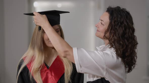 Smiling Mother Putting Graduation Cap Tassel Right to Left on Cap of Happy Daughter Standing Indoors alt