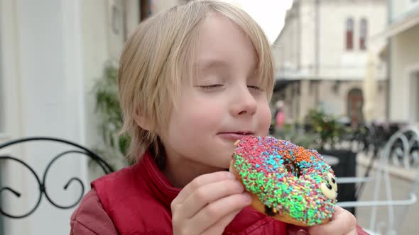 Cute school age boy eating funny colorful donut in a street cafe