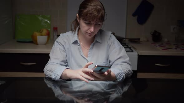 Serious Woman Sitting in the Kitchen at Night and Using the Phone