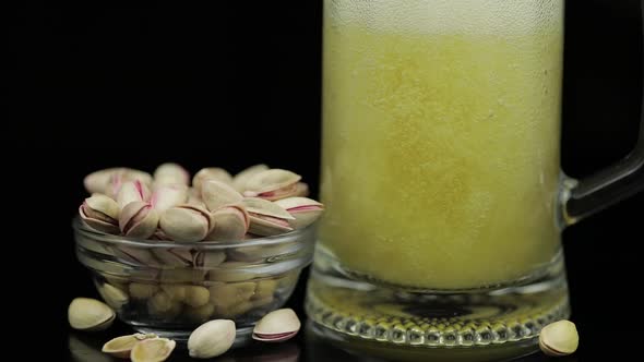 Beer Is Pouring Into Glass on Black Background. Bowl of Pistachios Nuts alt