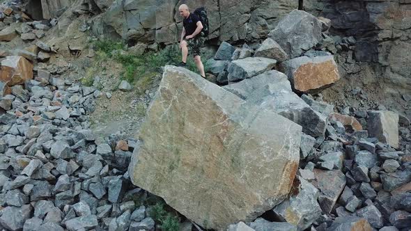 A man in a black t-shirt and a backpack comes to the edge of a large stone and raises his hands up. alt