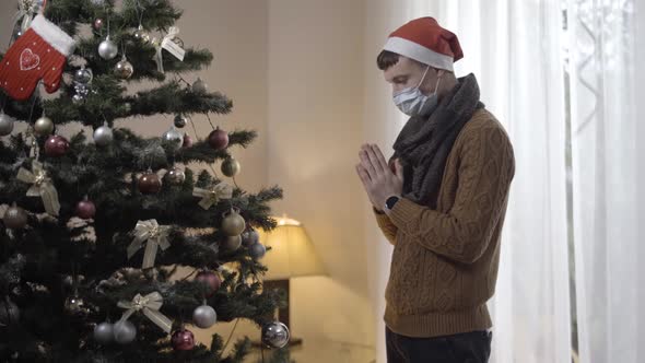 Side View of Hopeful Young Man in Covid19 Face Mask Praying To Christmas Tree at Home alt