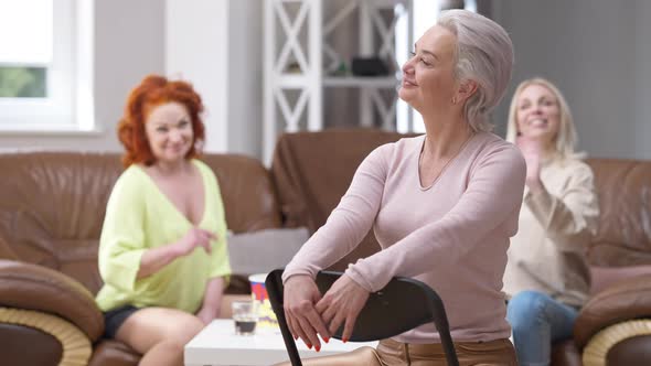 Confident Gorgeous Adult Woman Sitting on Chair at Home Looking Back at Friends Waving and Mocking alt