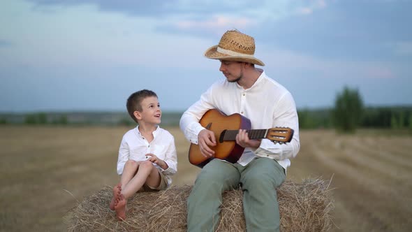 Little Boy and His Father Playing Guitar at Hay Bale in Field