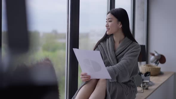 Happy Smiling Asian Female Freelancer in Bathrobe Looking Out the Window Sitting on Windowsill in alt